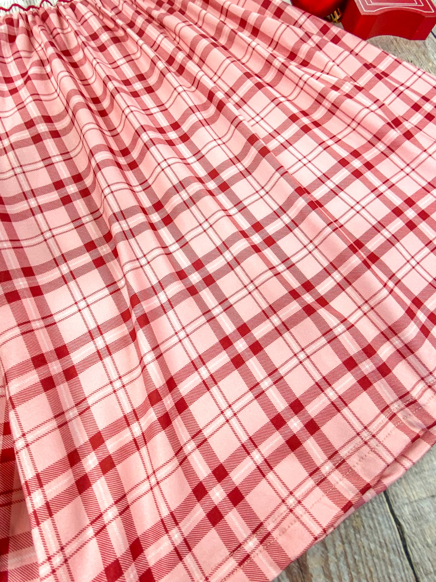 Close-up of the bottom hem of a red and pink plaid Christmas dress. The soft pleats and lightweight fabric are shown in detail, highlighting the gentle flow and texture of the dress against a wooden background.