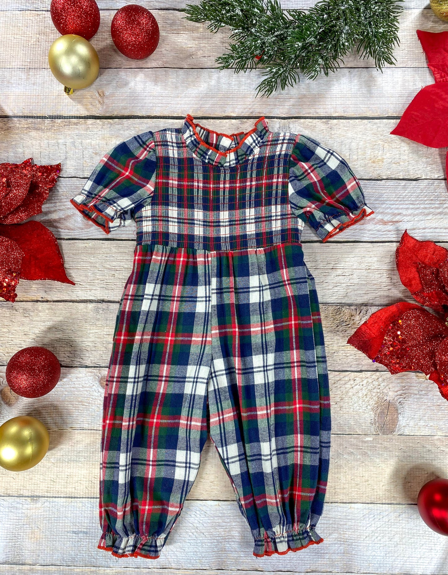 Flat lay of a girls’ plaid Christmas romper with a red, green, navy, and white checkered pattern. The romper has short puff sleeves, a ruffled mock neck, and elastic cuffs at the ankles with red trim. Surrounded by festive décor including red poinsettias, greenery, and gold and red ornaments.
