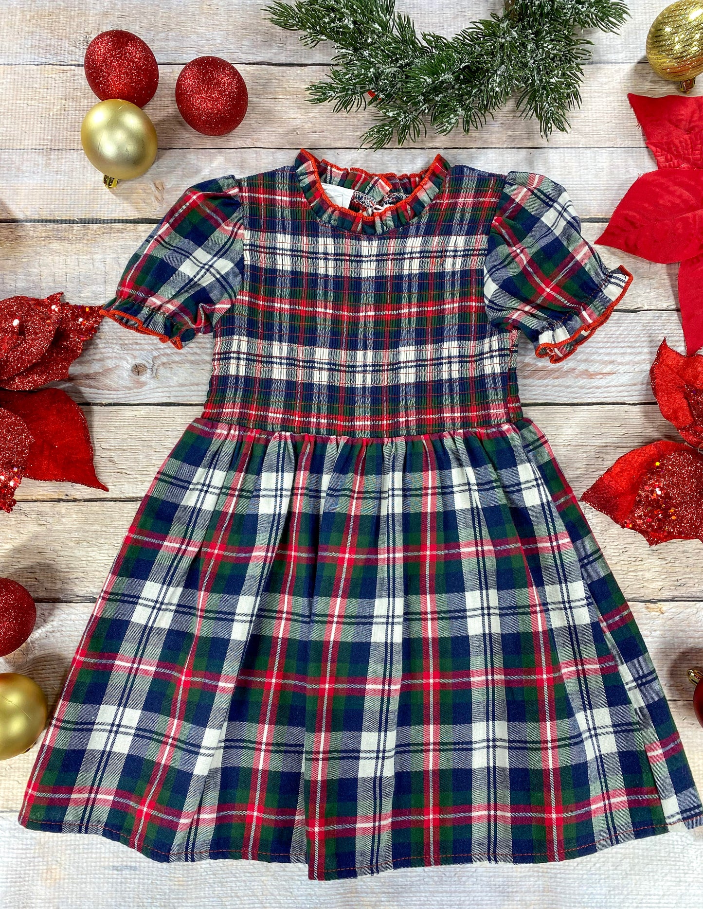 A classic holiday plaid dress for a young girl featuring red, green, navy, and white colors. The dress has puff sleeves, a gathered waist, and a ruffled collar trimmed in red. It’s displayed flat on a wood background surrounded by Christmas decorations, including red poinsettias, glittery red and gold ornaments, and evergreen foliage