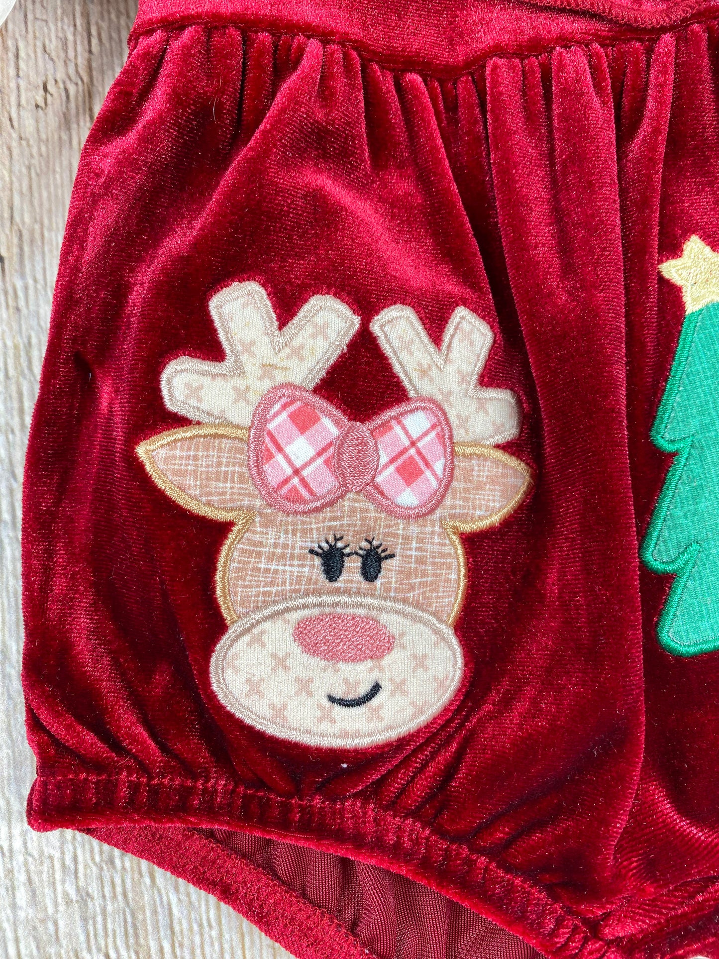 Zoomed-in image of the reindeer appliqué on the romper. The reindeer has a tan face with embroidered eyes and a smile, wearing red and white plaid bow and antlers. The texture of the fabric and embroidery detail are clearly visible