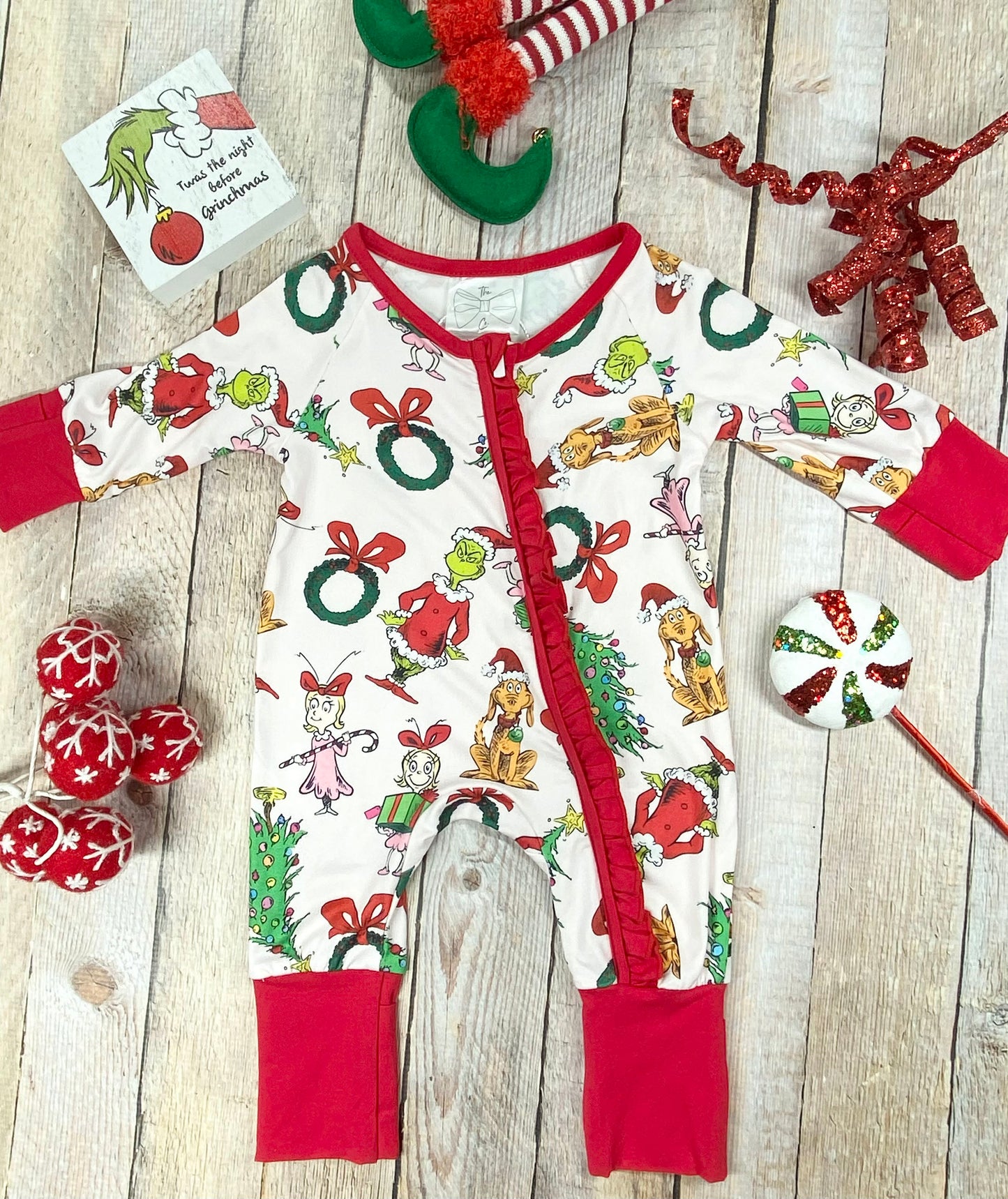 "Infant holiday romper with white fabric covered in playful Grinch characters, red bows, wreaths, and Christmas trees. Designed with vibrant red knit cuffs, a ruffled zipper front, and stretchy, soft material for cozy comfort."
