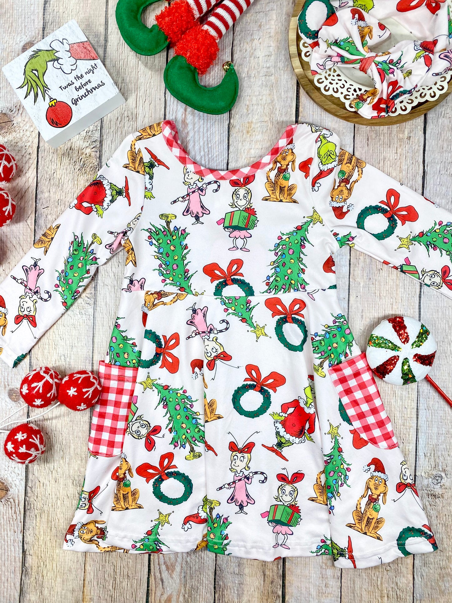 Girls’ long sleeve Christmas twirl dress featuring an all-over Grinch character print on white, with red and white gingham-trimmed pockets and matching neckline. Made from soft, stretchy knit fabric and cut for a twirl-friendly silhouette—perfect for holiday parties and photos.