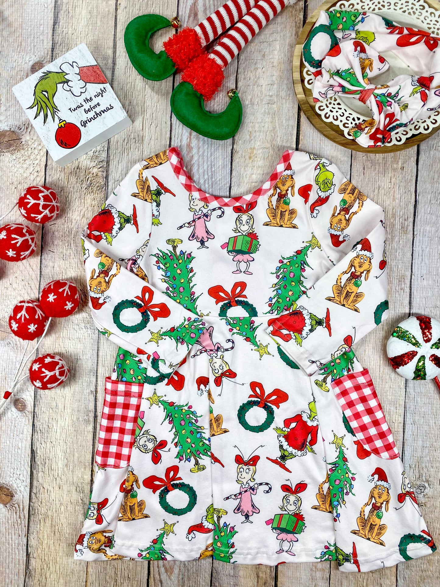 Girls’ long sleeve Christmas twirl dress featuring an all-over Grinch character print on white, with red and white gingham-trimmed pockets and matching neckline. Made from soft, stretchy knit fabric and cut for a twirl-friendly silhouette—perfect for holiday parties and photos.