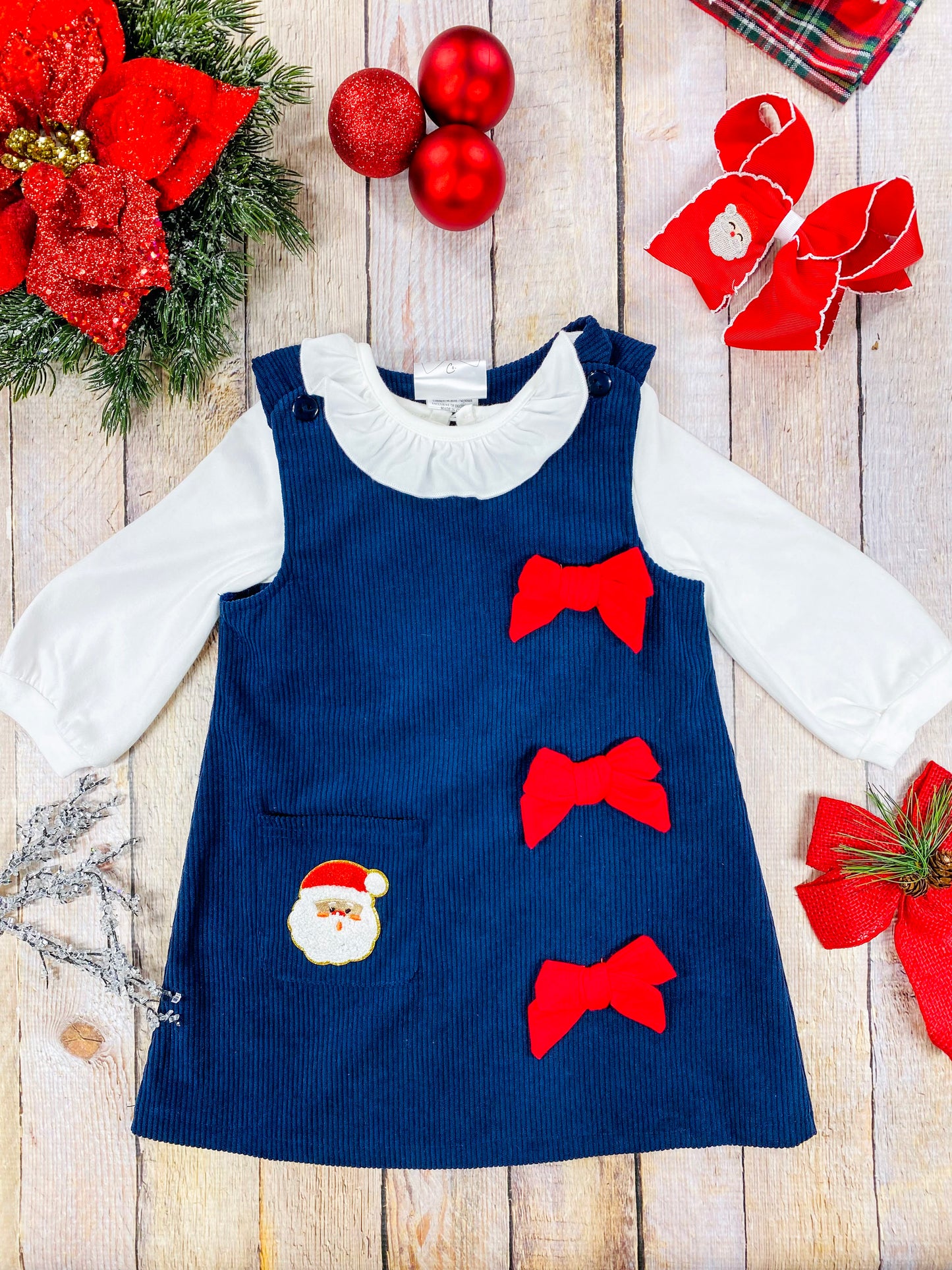 Toddler girls’ navy corduroy Christmas jumper dress featuring three red grosgrain bows on the right side and a Santa face appliqué on the front pocket. Layered over a white long sleeve ruffled blouse—perfect for classic holiday photos.