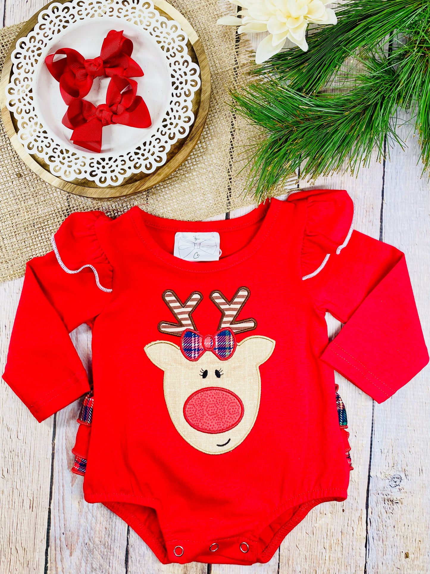 A red baby romper with long sleeves, ruffled shoulders trimmed in white, and a cute reindeer face appliqué on the front. The reindeer has plaid antlers, a red plaid bow, and a red nose. The outfit is displayed flat on a wooden background with pine branches, burlap fabric, and a white plate holding two red bows.