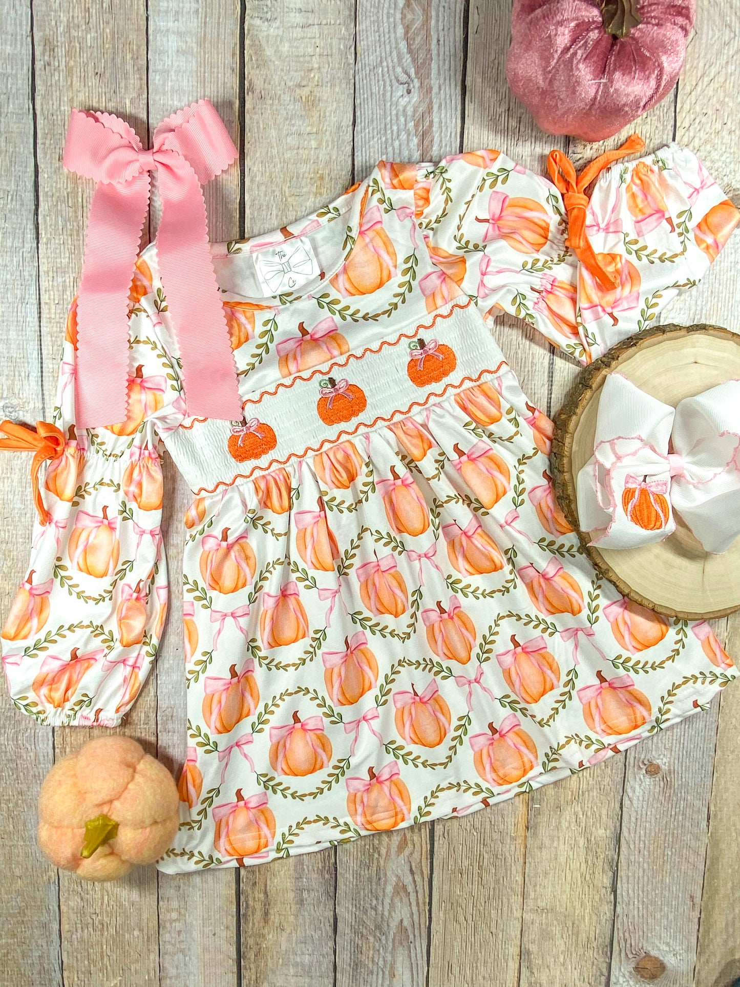 A long-sleeve dress with a white base and a pattern of orange pumpkins tied with pink bows, arranged within green leafy wreaths. The dress features smocking across the chest with embroidered orange pumpkins and pink bows. The sleeves have orange ribbon ties at the elbows. Beside the dress, there is a large pink hair bow with scalloped edges and a smaller white bow with a pumpkin embroidery placed on a wooden round slice (sold separately)