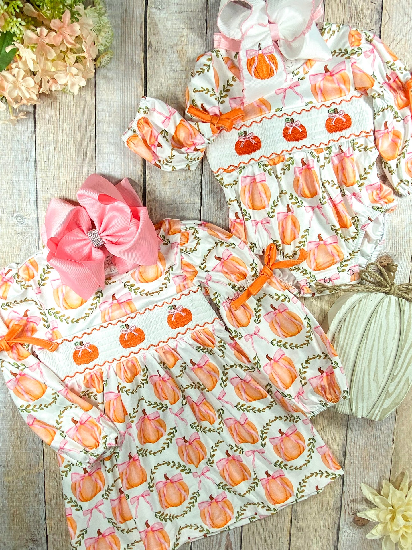 A flat lay display of two coordinating girls' fall outfits laid on a wooden surface. Both garments are decorated with a whimsical pumpkin print featuring orange pumpkins tied with pink bows and surrounded by green leafy wreaths. The top outfit is a long-sleeve romper with orange ribbon ties at the sleeves and a smocked chest panel embroidered with three orange pumpkins. It is accessorized with a large white hair bow featuring an embroidered pumpkin. The bottom outfit is a matching long-sleeve dress with the