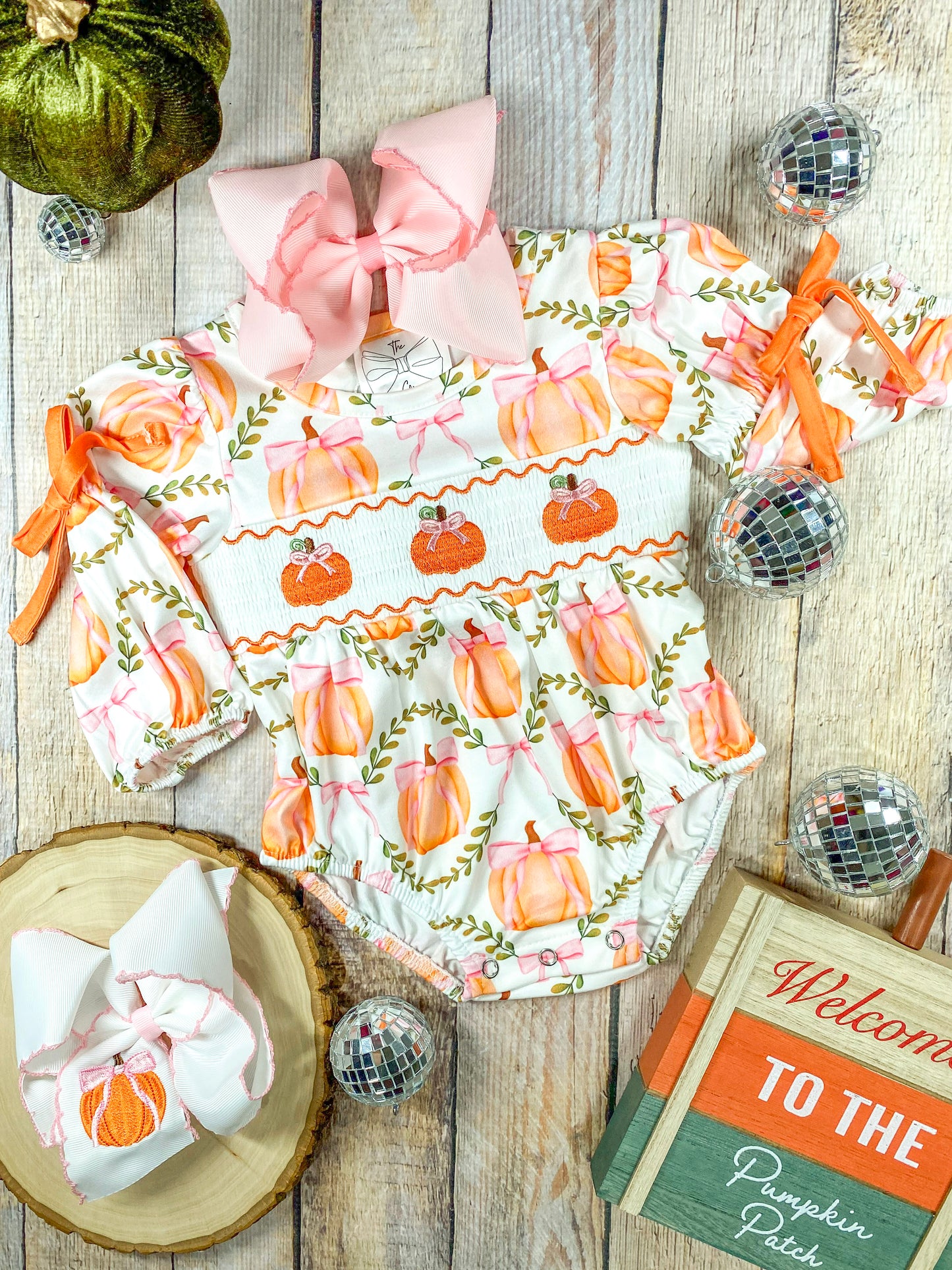 A flat lay of a infant girl's autumn-themed romper arranged on a wooden surface. The outfit features a long-sleeved romper decorated with pink and orange pumpkins tied with pink bows and green vines. The middle section has smocking with embroidered orange pumpkins wearing pink bows. The sleeves are gathered with orange ribbon ties. A large pink hair bow is placed at the neckline, and a matching white hair bow with a pumpkin embroidery rests on a wooden slice to the left (both bows sold separately)
