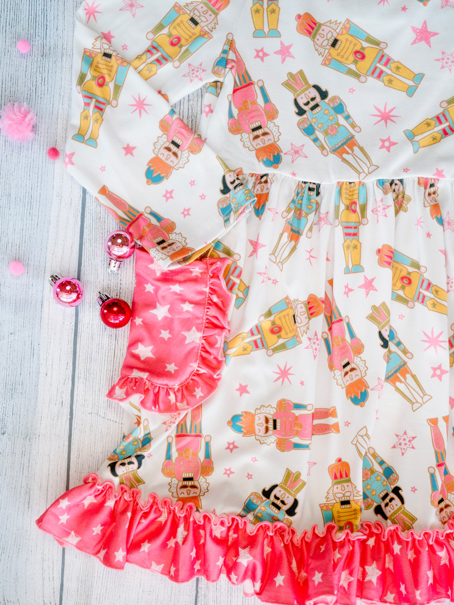 Close-up of the Nutcracker Galore Twirl Dress with Pockets, featuring a colorful nutcracker print on white fabric with pink star accents. The dress includes pink star-patterned side pockets with ruffled trim and a coordinating ruffle hem along the skirt.