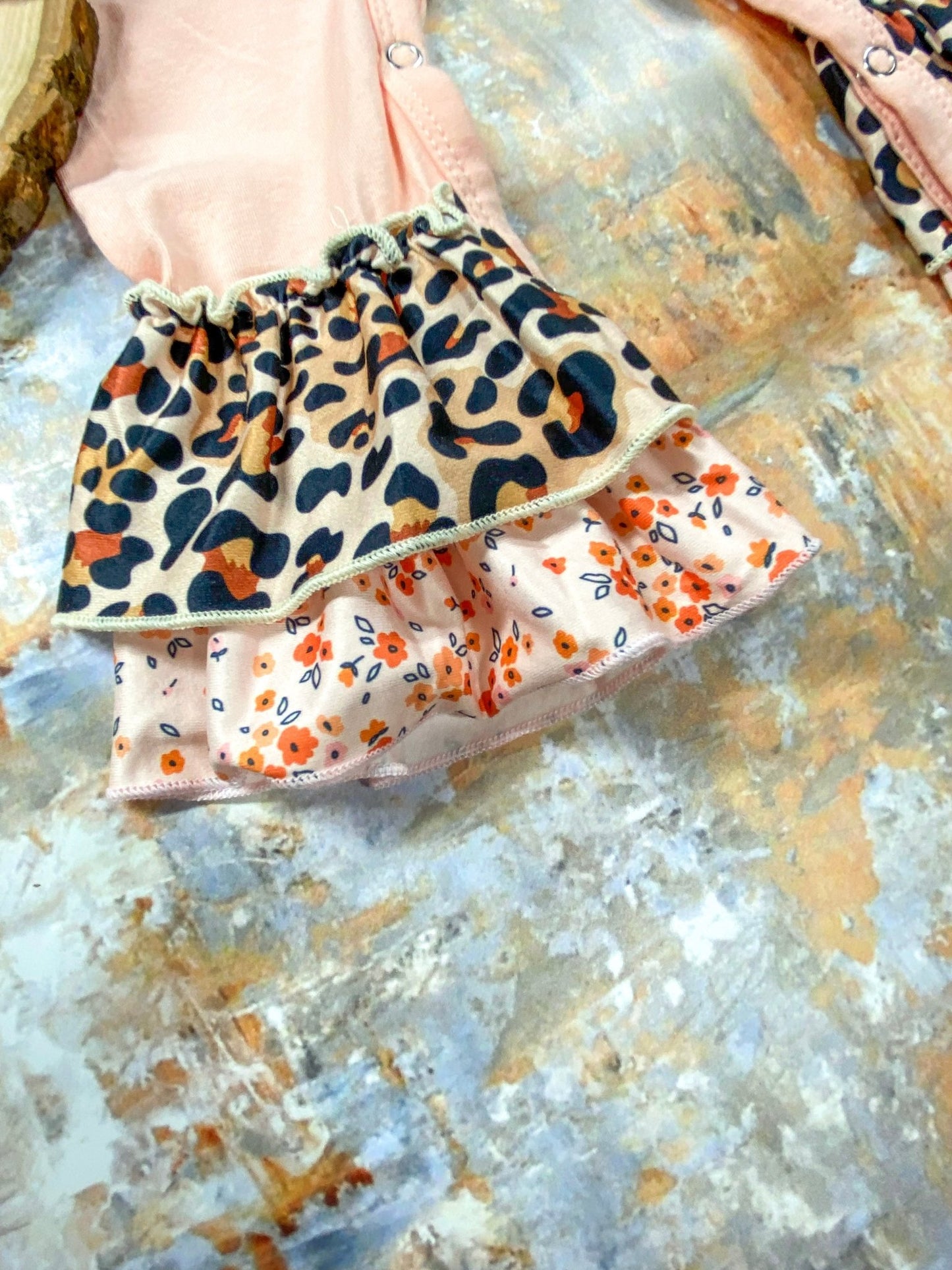 Close-up of the leg cuff on a pink baby romper featuring double-layered ruffles—top layer in a tan leopard print and bottom layer in a peach floral pattern with small orange flowers and blue leaves, displayed on a textured surface.