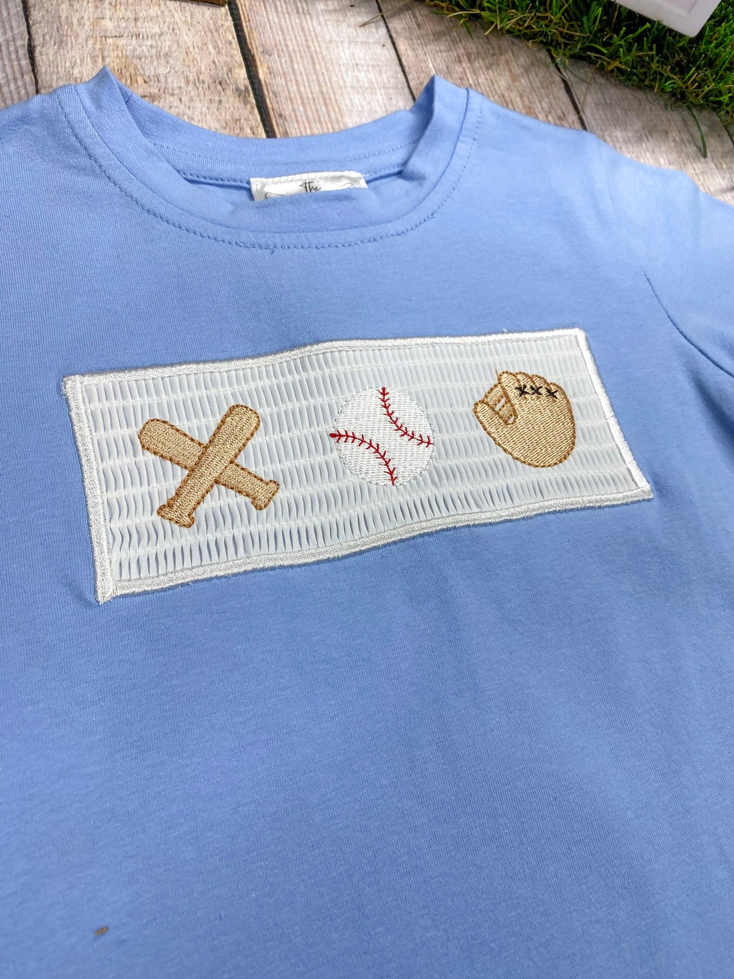 Blue shirt with sports-themed patch featuring a baseball bat, ball, and glove on a wooden surface.