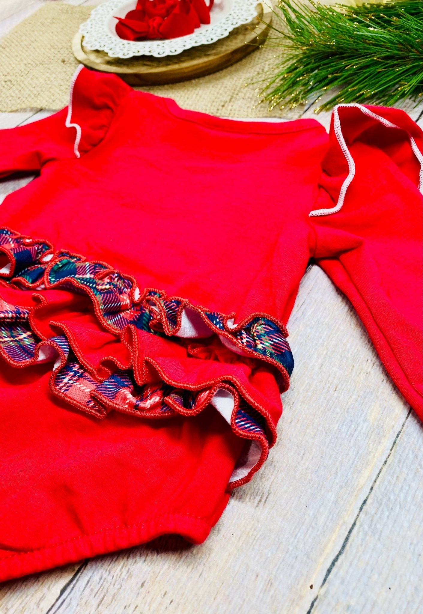 close up of the back of a festive red baby romper with long sleeves and white trim along ruffled shoulders. The bottom half features two rows of red and plaid ruffles across the back.