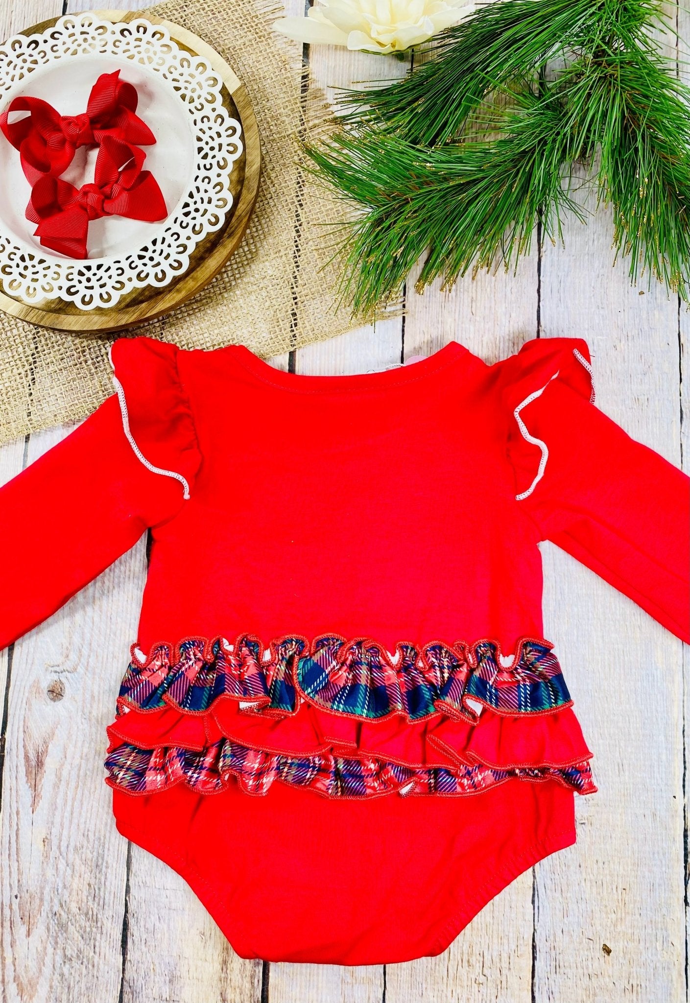 The back of a festive red baby romper with long sleeves and white trim along ruffled shoulders. The bottom half features two rows of red and plaid ruffles across the back. It’s laid flat on a rustic wooden surface beside a white decorative plate with two red bows, burlap fabric, green pine branches, and a white flower.
