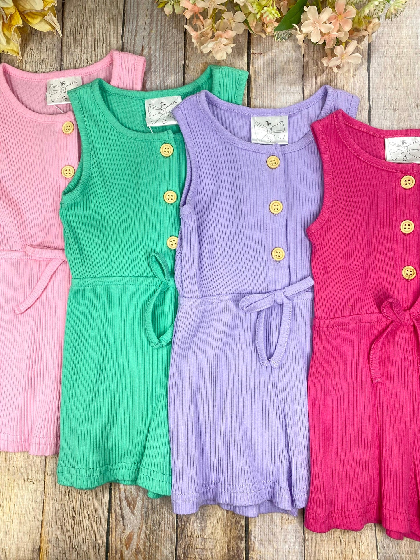 Four colorful ribbed tank dresses on a wooden surface with flowers in the background.