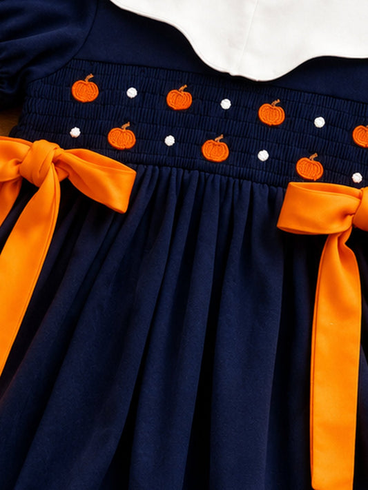 Close-up of orange pumpkin smocking, white accents and orange waist bows on a navy girls fall dress. - Alternative view
