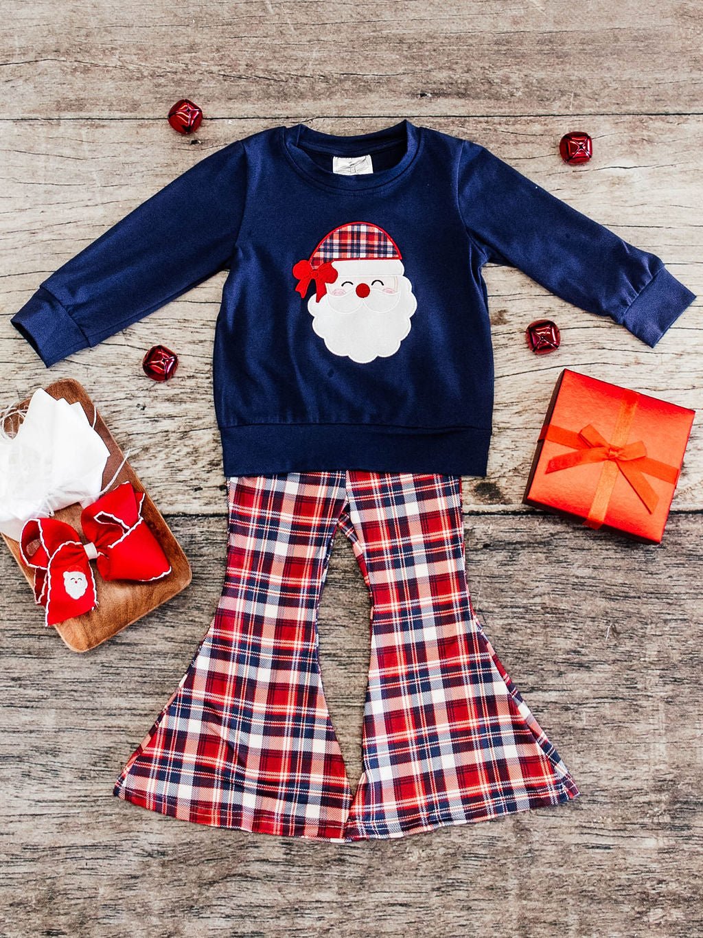 flat lay of an outfit featuring a navy long sleeve with a santa wearing a plaid hat applique in the center and paired with red, navy, and white plaid bell botto pants against a wooden background with ornament decorations, a red gift box, and red bow with a santa embroidery on it