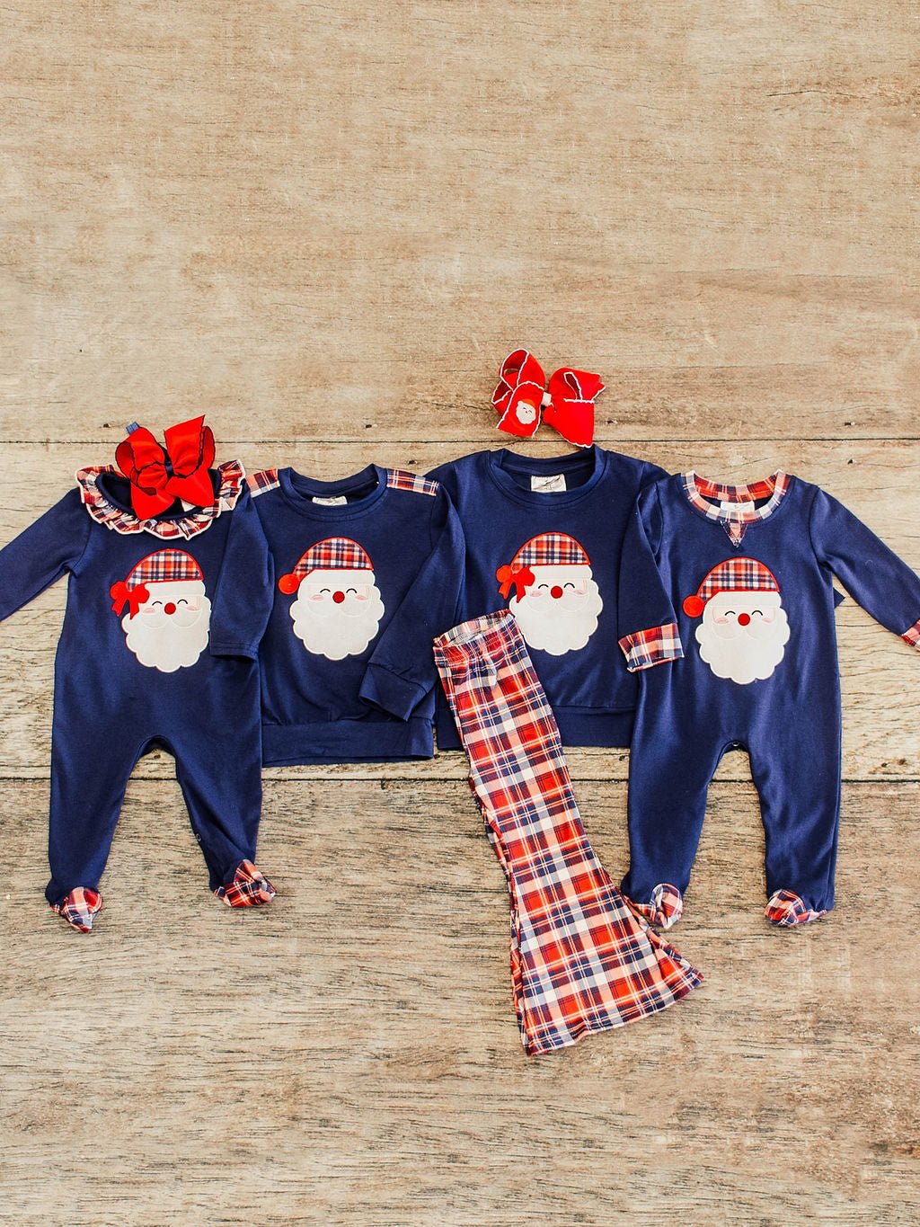 A coordinated family Christmas outfit set laid out on a wooden background. The collection includes two navy blue baby rompers and two long-sleeve shirts, all featuring a Santa Claus face with a red plaid hat and bow. Matching red plaid pants are placed in the center. The baby rompers have plaid cuffs, and accessories include a large red bow headband
