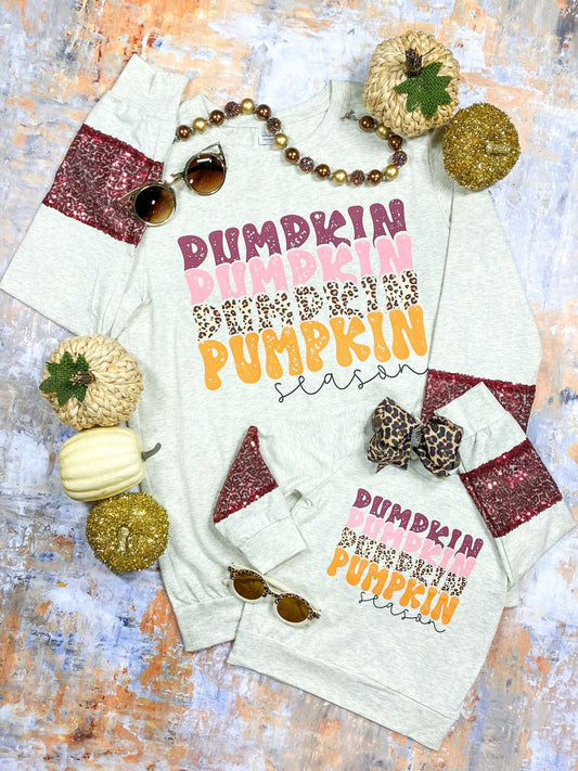 Matching fall sweatshirts in adult and child sizes with maroon sequin sleeve accents. Both sweatshirts are light gray and feature the text “Pumpkin Season” with stacked designs in maroon, pink, leopard print, and orange. Styled flat lay with beaded necklace, sunglasses, leopard print bow, and decorative pumpkins in cream, gold, and natural tones - Alternative view