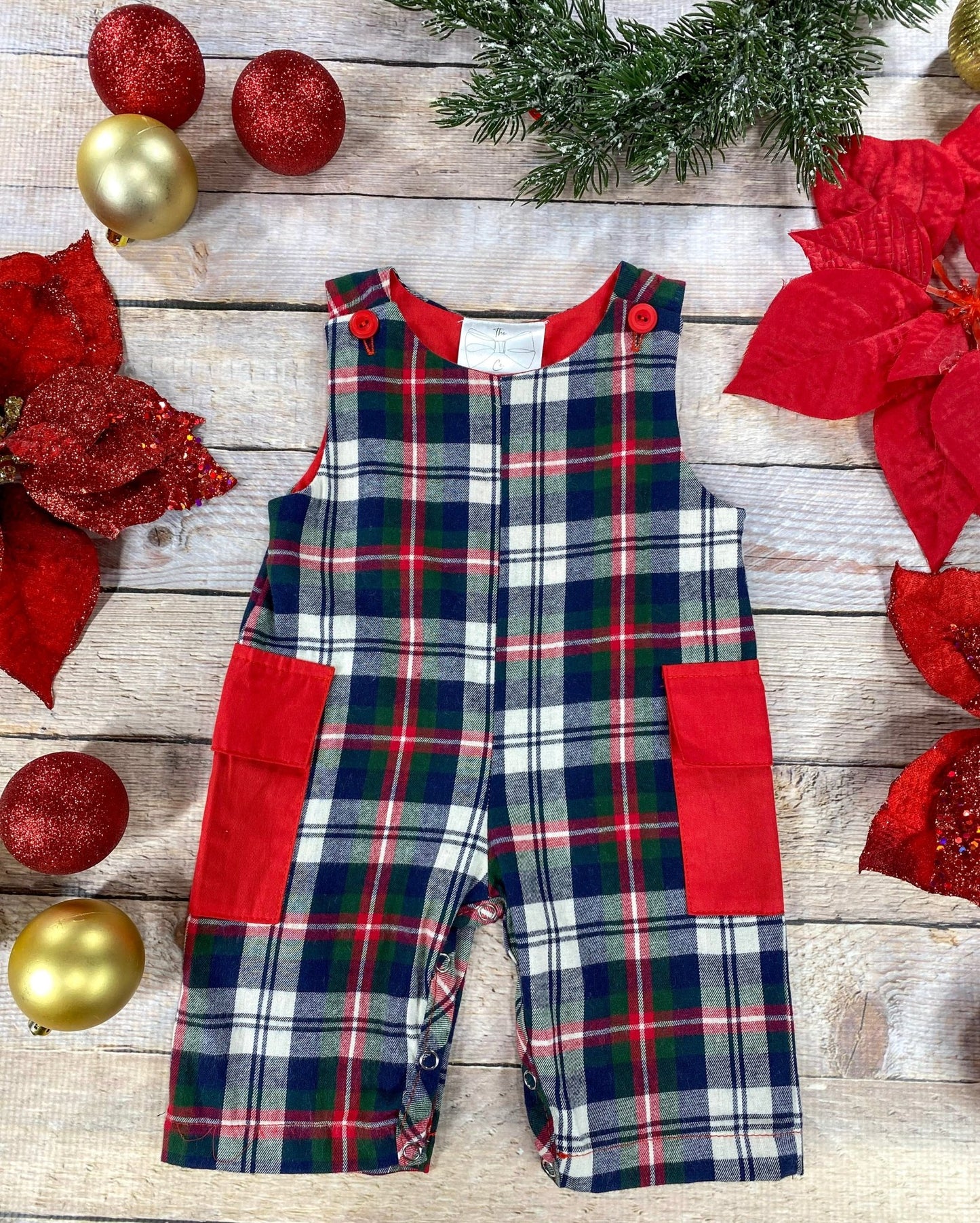 Flat lay of boys’ Christmas plaid overalls in red, green, navy, and white. The overalls feature red shoulder buttons and red patch pockets on the sides, with snap closures along the inseam for easy changes. Styled with gold and red ornaments, poinsettias, and greenery on a light wood background.