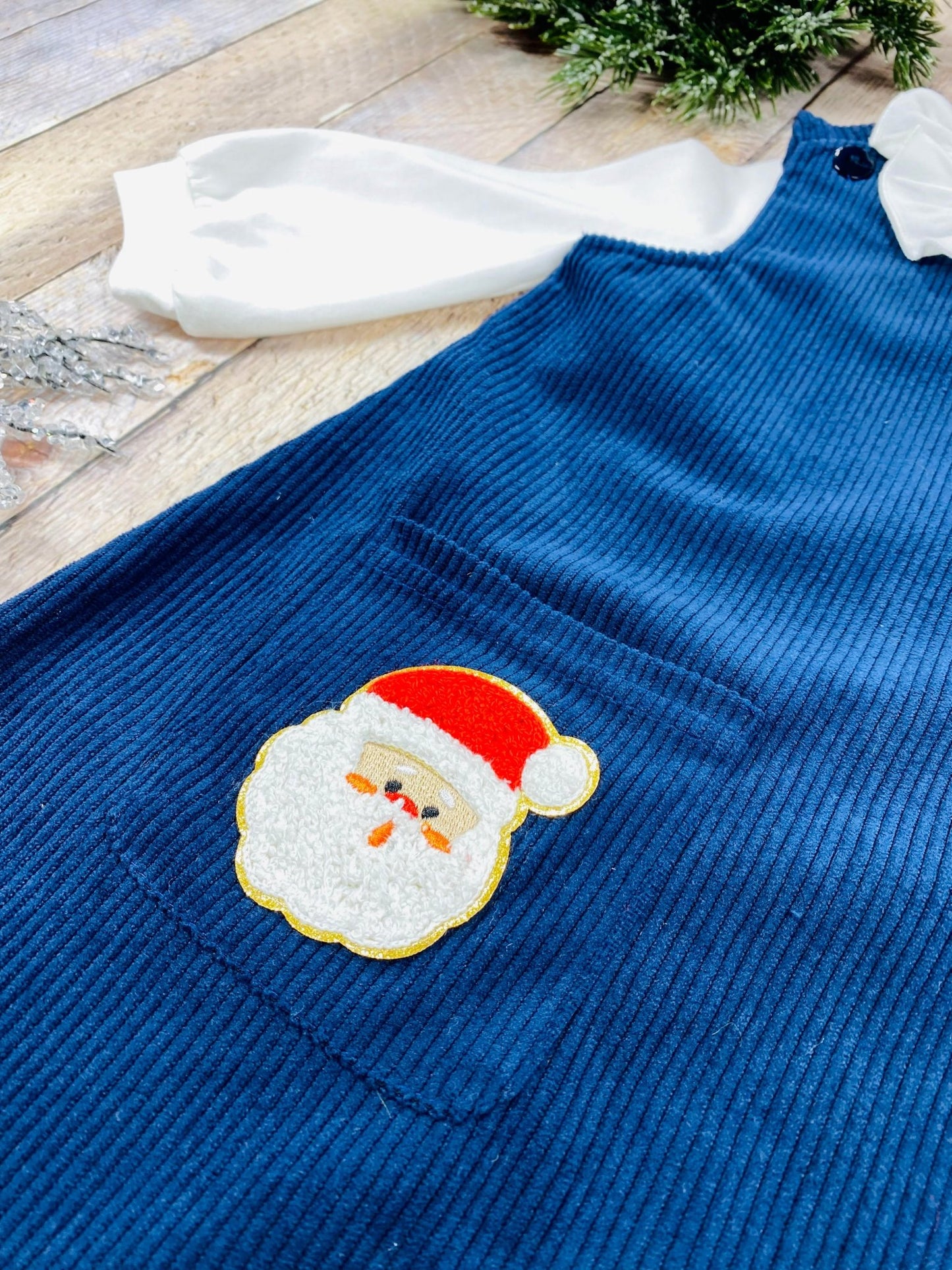 Detailed close-up of embroidered Santa Claus appliqué on navy blue corduroy pocket, featuring classic Santa face with white beard, red hat, and cheerful expression—adding a festive and playful holiday touch to the outfit.