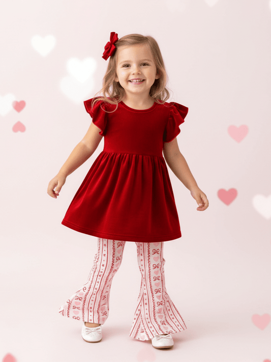 Young girl in a red top with ruffled sleeves and patterned leggings standing against a light pink background with heart shapes. - Alternative view