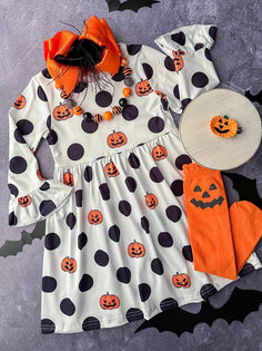 Flat lay of a girl's Halloween outfit featuring a white dress with black polka dots and jack-o’-lantern prints, orange pumpkin tights, a chunky orange and black beaded necklace, a large orange bow with feathers, and a pumpkin hair clip.