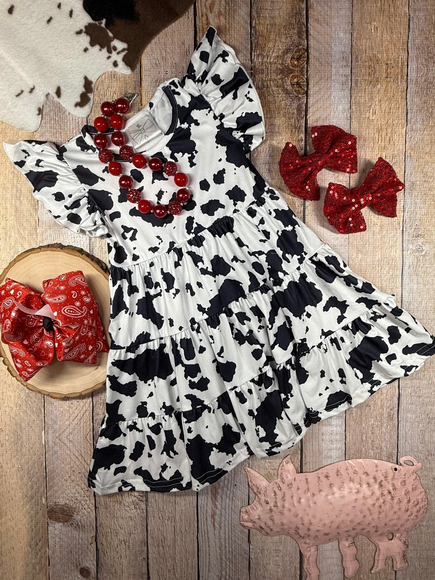 Flat lay of a toddler fall outfit with an orange knit sweater, pumpkin-print skirt, black fringe bag, black and white beaded necklaces, white and orange bows, black bow, and round sunglasses on a pink backgrou