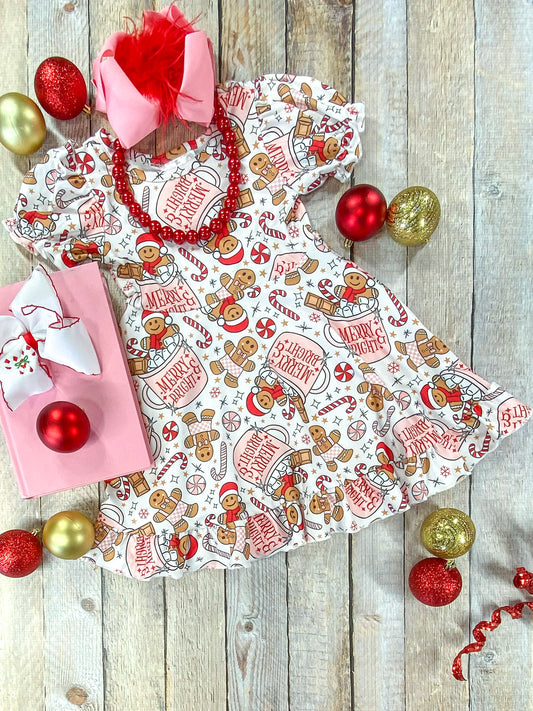 Full flat lay of a girls’ Christmas dress with puff sleeves and a playful holiday print of gingerbread cookies, candy canes, hot cocoa mugs reading “Merry & Bright,” and snowflakes on a cream background, styled with a pink bow, red bead necklace, gift box, and festive ornaments.