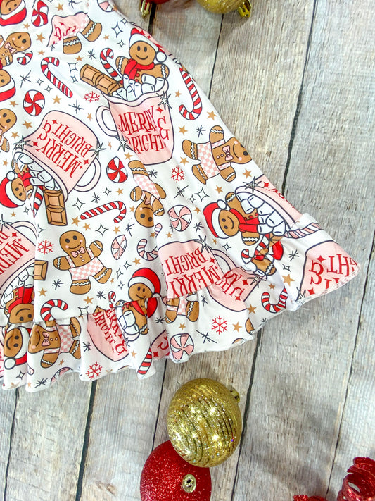 Detailed view of the ruffled hem of a holiday girls’ dress decorated with cheerful gingerbread figures, candy canes, snowflakes, and “Merry & Bright” cocoa mugs, styled with glittery red and gold Christmas ornaments on a wood background. - Alternative view