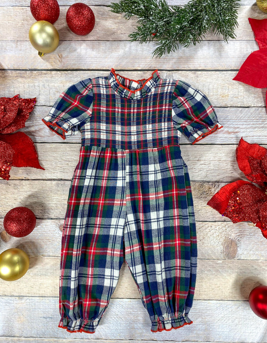 Flat lay of a girls’ plaid Christmas romper with a red, green, navy, and white checkered pattern. The romper has short puff sleeves, a ruffled mock neck, and elastic cuffs at the ankles with red trim. Surrounded by festive décor including red poinsettias, greenery, and gold and red ornaments.