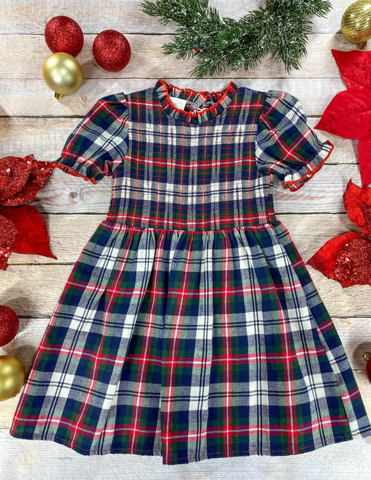 A classic holiday plaid dress for a young girl featuring red, green, navy, and white colors. The dress has puff sleeves, a gathered waist, and a ruffled collar trimmed in red. It’s displayed flat on a wood background surrounded by Christmas decorations, including red poinsettias, glittery red and gold ornaments, and evergreen foliage