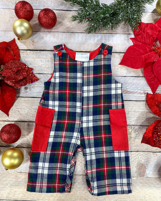 Flat lay of boys’ Christmas plaid overalls in red, green, navy, and white. The overalls feature red shoulder buttons and red patch pockets on the sides, with snap closures along the inseam for easy changes. Styled with gold and red ornaments, poinsettias, and greenery on a light wood background.