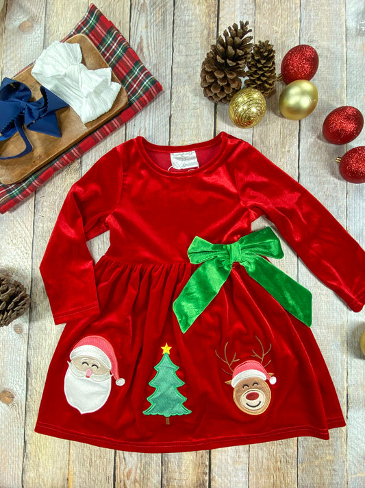 A toddler girl's red velvet holiday dress with long sleeves and a large green velvet bow at the waist. The skirt is decorated with cheerful Christmas appliqués: Santa, a Christmas tree, and a smiling reindeer with antlers and a Santa hat. The dress is laid out on a rustic wooden background, surrounded by pinecones, gold and red ornaments, and plaid fabric, with a tray holding large navy and white hair bows
