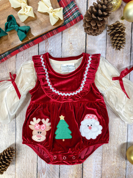 A festive red velvet baby romper with cream sheer long sleeves tied with red ribbons. The front features three Christmas-themed appliqués: a reindeer with a plaid bow, a green Christmas tree, and a Santa face. The romper has frilly ruffle detailing around the chest and buttons at the bottom for easy diaper changes. It’s displayed on a wooden background with pinecones, gold ornaments, and plaid fabric, along with a tray holding green and cream hair bows