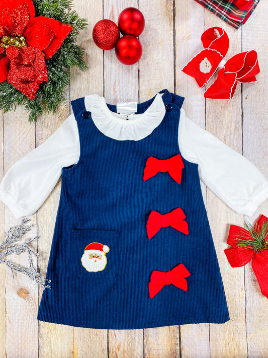 Toddler girls’ navy corduroy Christmas jumper dress featuring three red grosgrain bows on the right side and a Santa face appliqué on the front pocket. Layered over a white long sleeve ruffled blouse—perfect for classic holiday photos.