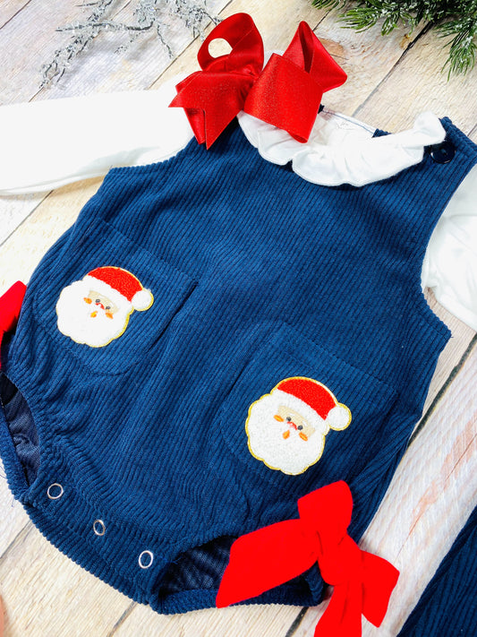 Infant girls’ navy blue corduroy romper with long white puff sleeves, red grosgrain bows at the hips, and front patch pockets with embroidered Santa Claus appliqués. Snap closure at the bottom for easy changes.