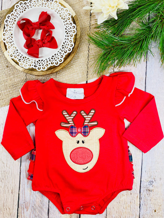 A red baby romper with long sleeves, ruffled shoulders trimmed in white, and a cute reindeer face appliqué on the front. The reindeer has plaid antlers, a red plaid bow, and a red nose. The outfit is displayed flat on a wooden background with pine branches, burlap fabric, and a white plate holding two red bows.