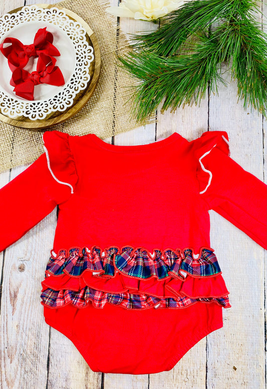 The back of a festive red baby romper with long sleeves and white trim along ruffled shoulders. The bottom half features two rows of red and plaid ruffles across the back. It’s laid flat on a rustic wooden surface beside a white decorative plate with two red bows, burlap fabric, green pine branches, and a white flower. - Alternative view