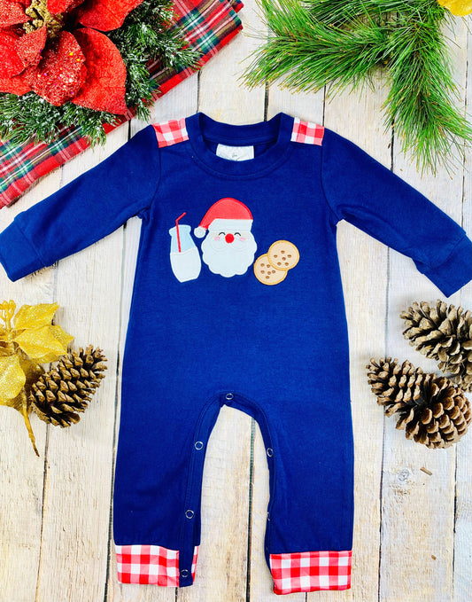 A navy blue baby romper with long sleeves and plaid red-and-white cuffs at the ankles and shoulders. The front features an appliqué of Santa’s face next to a glass of milk and two cookies. The outfit is laid flat on a light wooden background decorated with pinecones, red plaid fabric, a poinsettia, and evergreen branches for a holiday theme.
