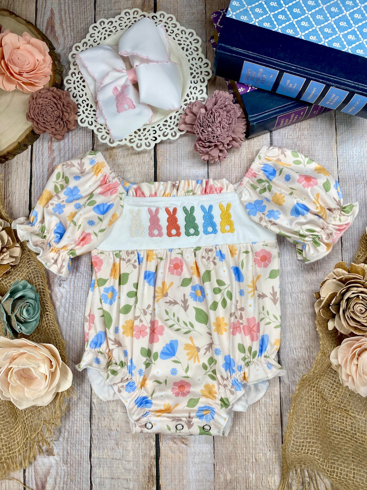 Colorful baby romper with floral and bunny designs on a wooden surface with decorative flowers.