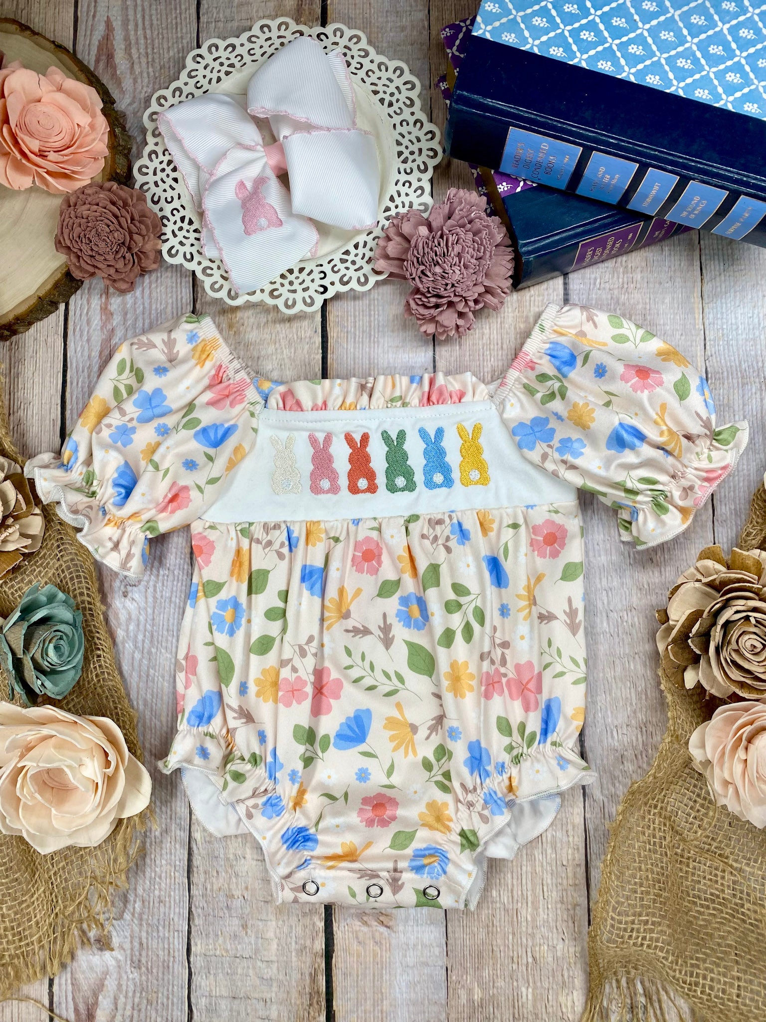 Colorful baby romper with floral and bunny designs on a wooden surface with decorative flowers.