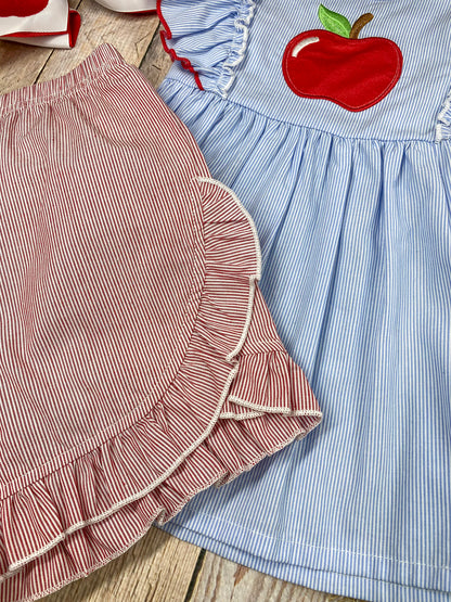 Children's outfit with blue top and pink shorts featuring an apple design on a wooden background