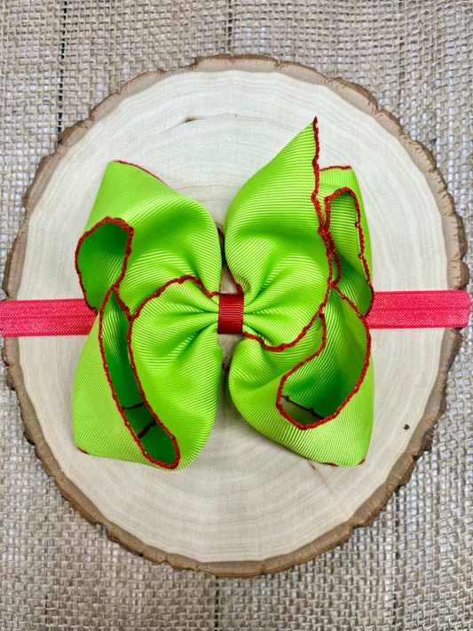 Green hair bow with red accents on a pink headband, placed on a wooden surface.