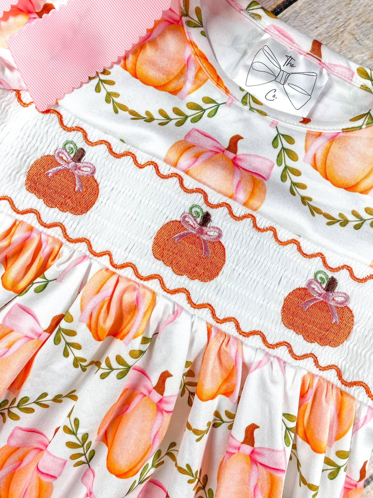 A close-up view of a white fabric dress with an embroidered design featuring three orange pumpkins with pink bows on the chest area. The dress also has a pattern of larger pumpkins with pink bows and green leafy vines on the main fabric.