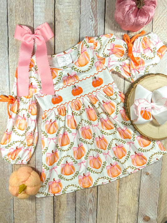A long-sleeve dress with a white base and a pattern of orange pumpkins tied with pink bows, arranged within green leafy wreaths. The dress features smocking across the chest with embroidered orange pumpkins and pink bows. The sleeves have orange ribbon ties at the elbows. Beside the dress, there is a large pink hair bow with scalloped edges and a smaller white bow with a pumpkin embroidery placed on a wooden round slice (sold separately)
