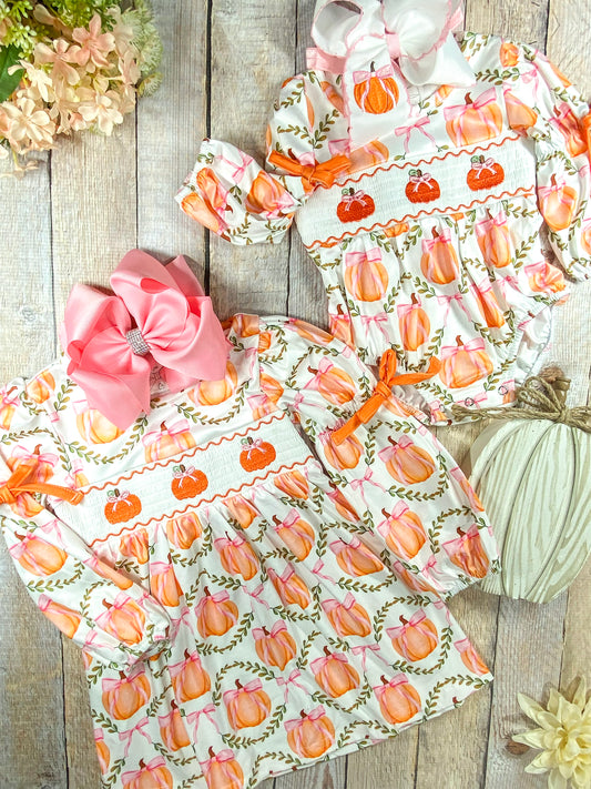 A flat lay display of two coordinating girls' fall outfits laid on a wooden surface. Both garments are decorated with a whimsical pumpkin print featuring orange pumpkins tied with pink bows and surrounded by green leafy wreaths. The top outfit is a long-sleeve romper with orange ribbon ties at the sleeves and a smocked chest panel embroidered with three orange pumpkins. It is accessorized with a large white hair bow featuring an embroidered pumpkin. The bottom outfit is a matching long-sleeve dress with the - Alternative view