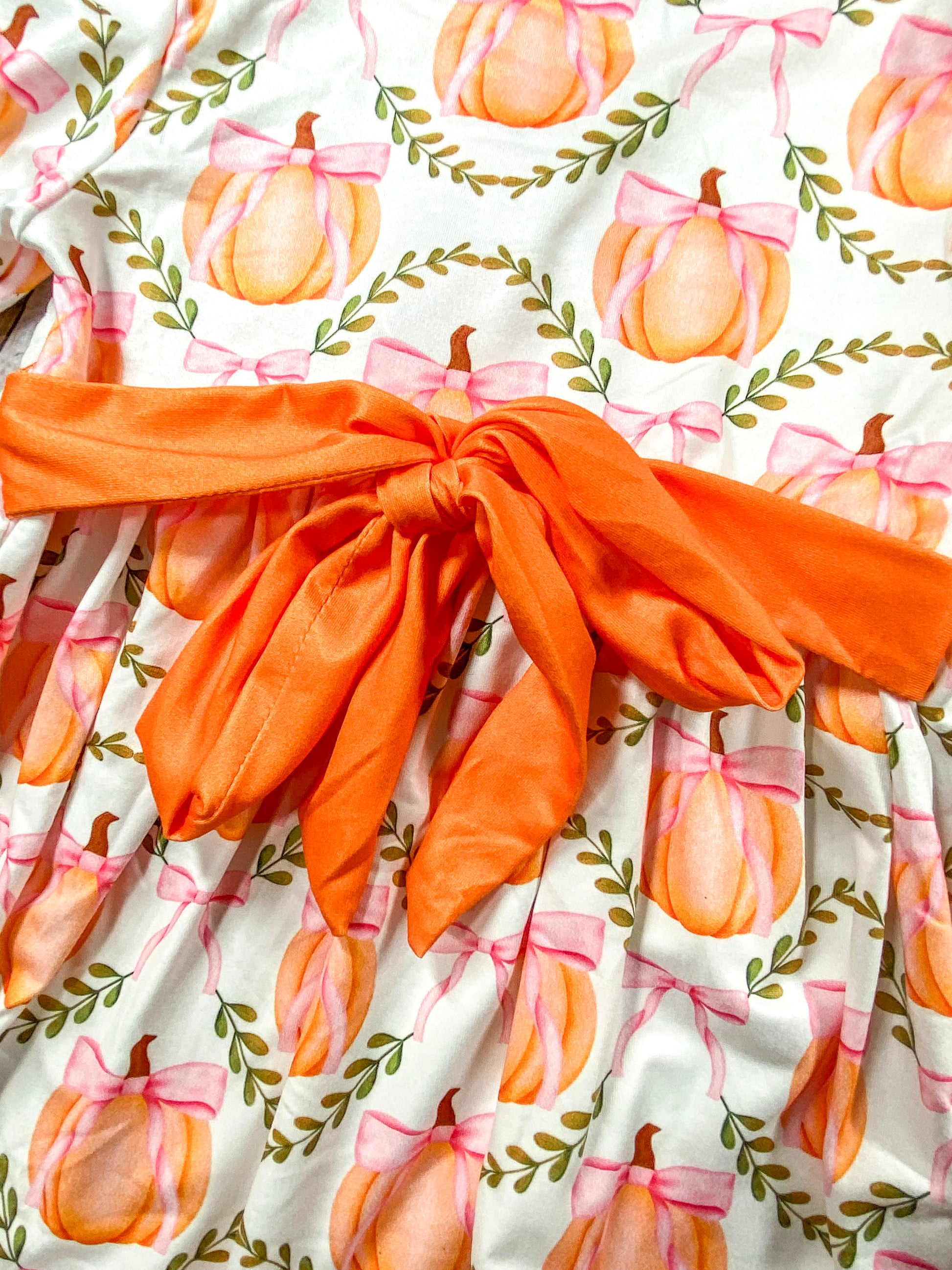 A close-up of a fabric with a pattern of orange pumpkins tied with pink bows, surrounded by green leafy vines. The fabric also features a prominent large orange bow tied at the center.