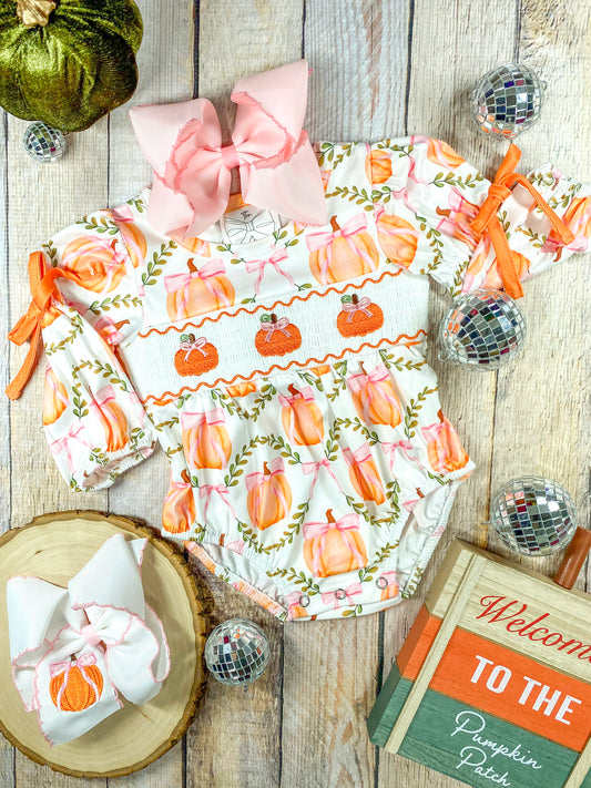 A flat lay of a infant girl's autumn-themed romper arranged on a wooden surface. The outfit features a long-sleeved romper decorated with pink and orange pumpkins tied with pink bows and green vines. The middle section has smocking with embroidered orange pumpkins wearing pink bows. The sleeves are gathered with orange ribbon ties. A large pink hair bow is placed at the neckline, and a matching white hair bow with a pumpkin embroidery rests on a wooden slice to the left (both bows sold separately)