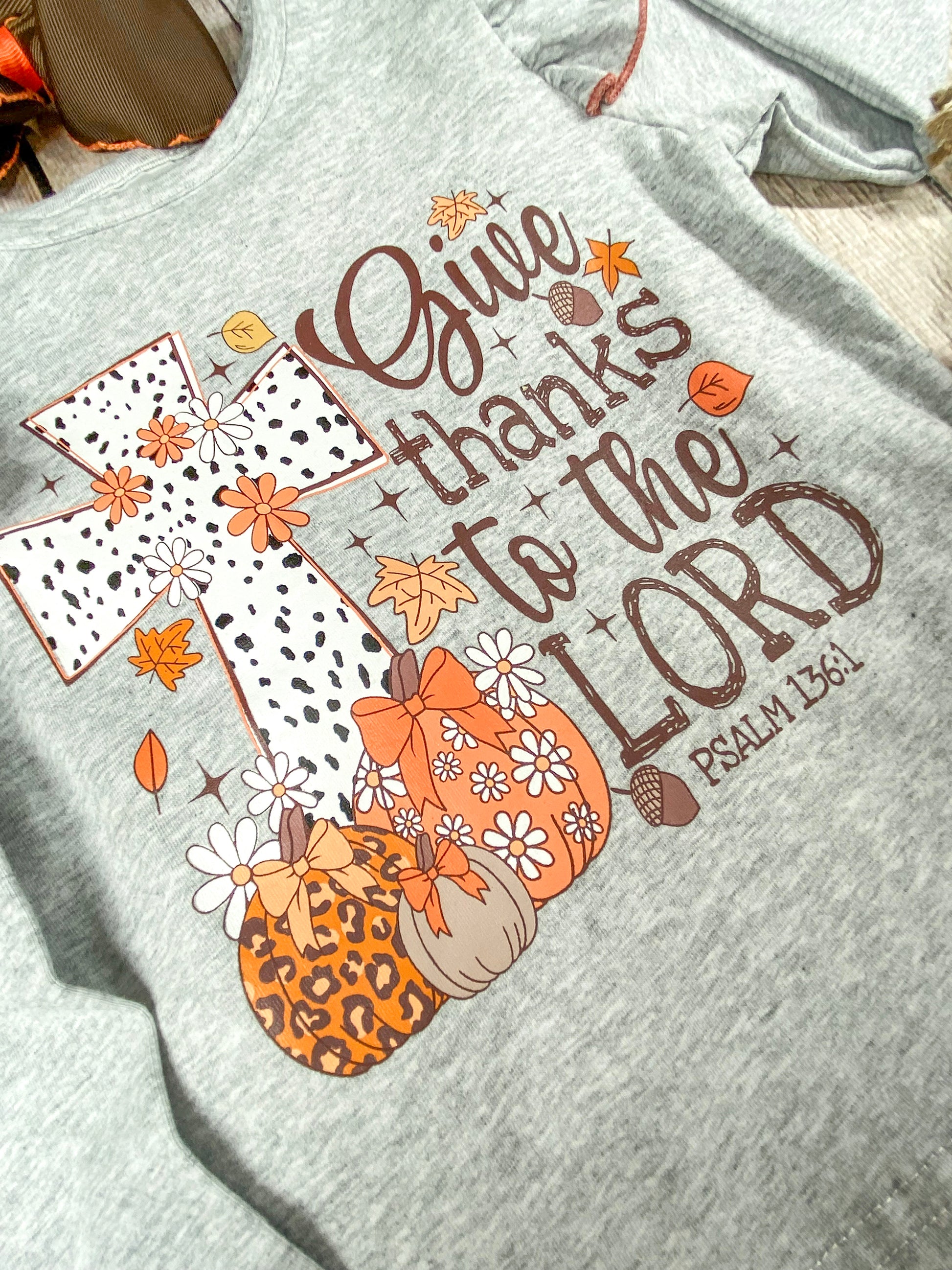 close up of the print text saying "Give thanks to the Lord Psalm 136:1" with a cross with some pumpkins in front of the cross on grey long sleeve