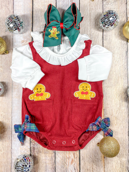 red corduroy romper with 2 gingerbread men appliques sticking out of pcokets on each side with a white long sleeve with ruffled collar layered underneath it and 2 plaid bows near leg openings